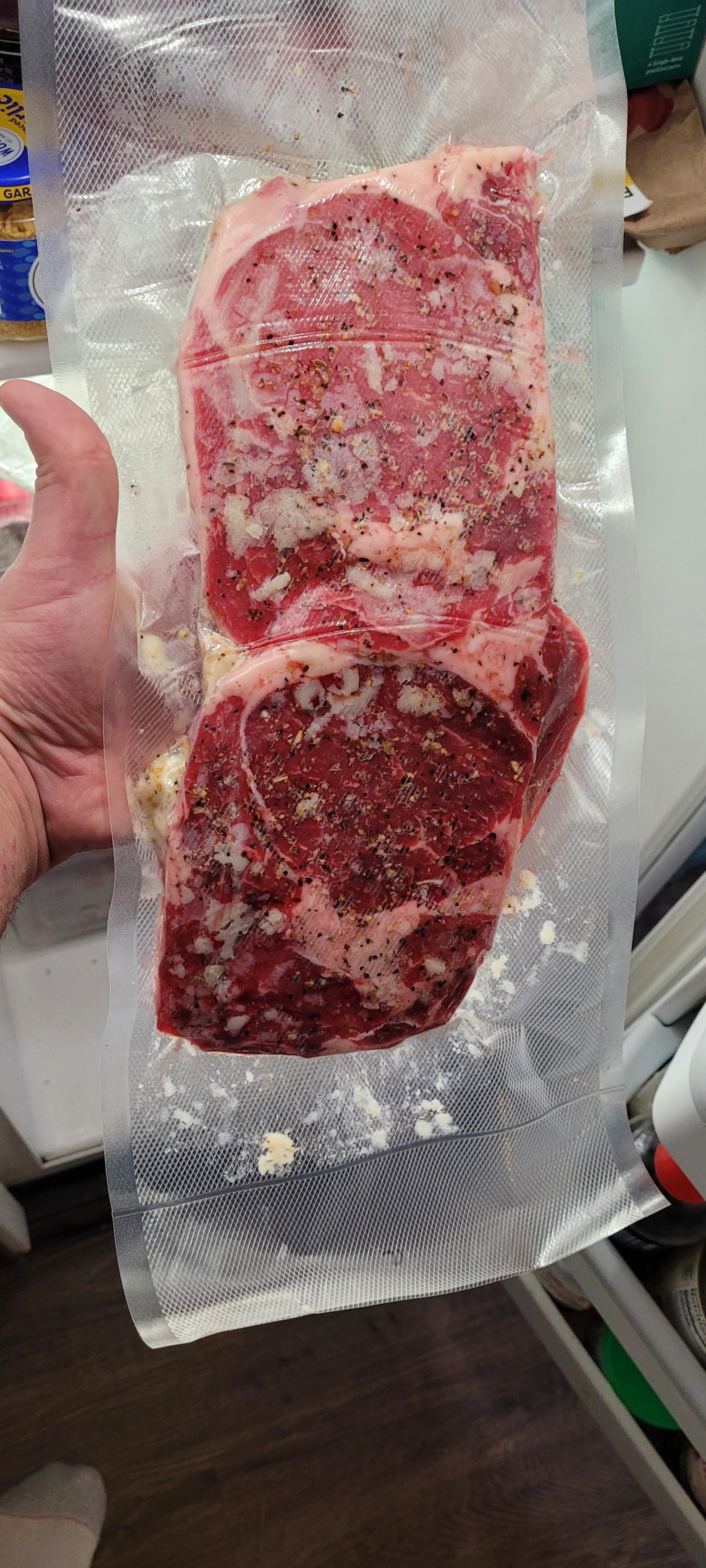 A vacuum-sealed package with two raw seasoned steaks being held by a hand over a kitchen counter