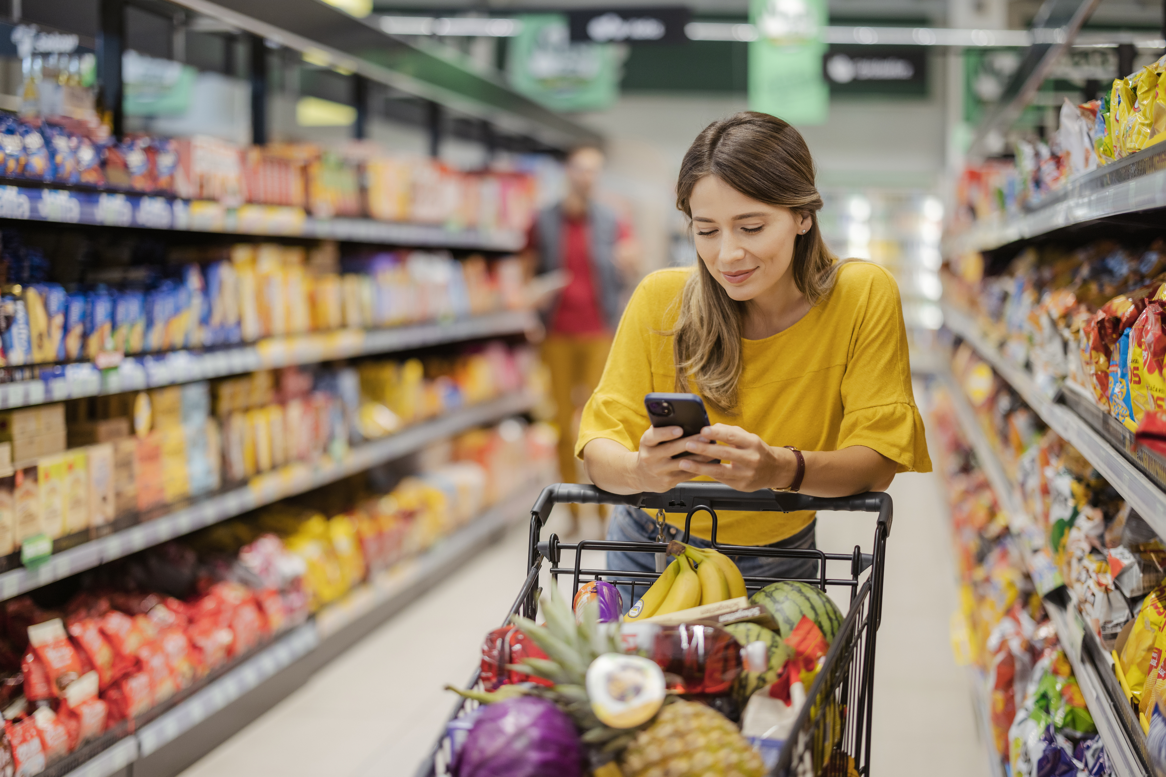 A person is shopping in a grocery store, pushing a cart of various fruits and vegetables while looking at their phone