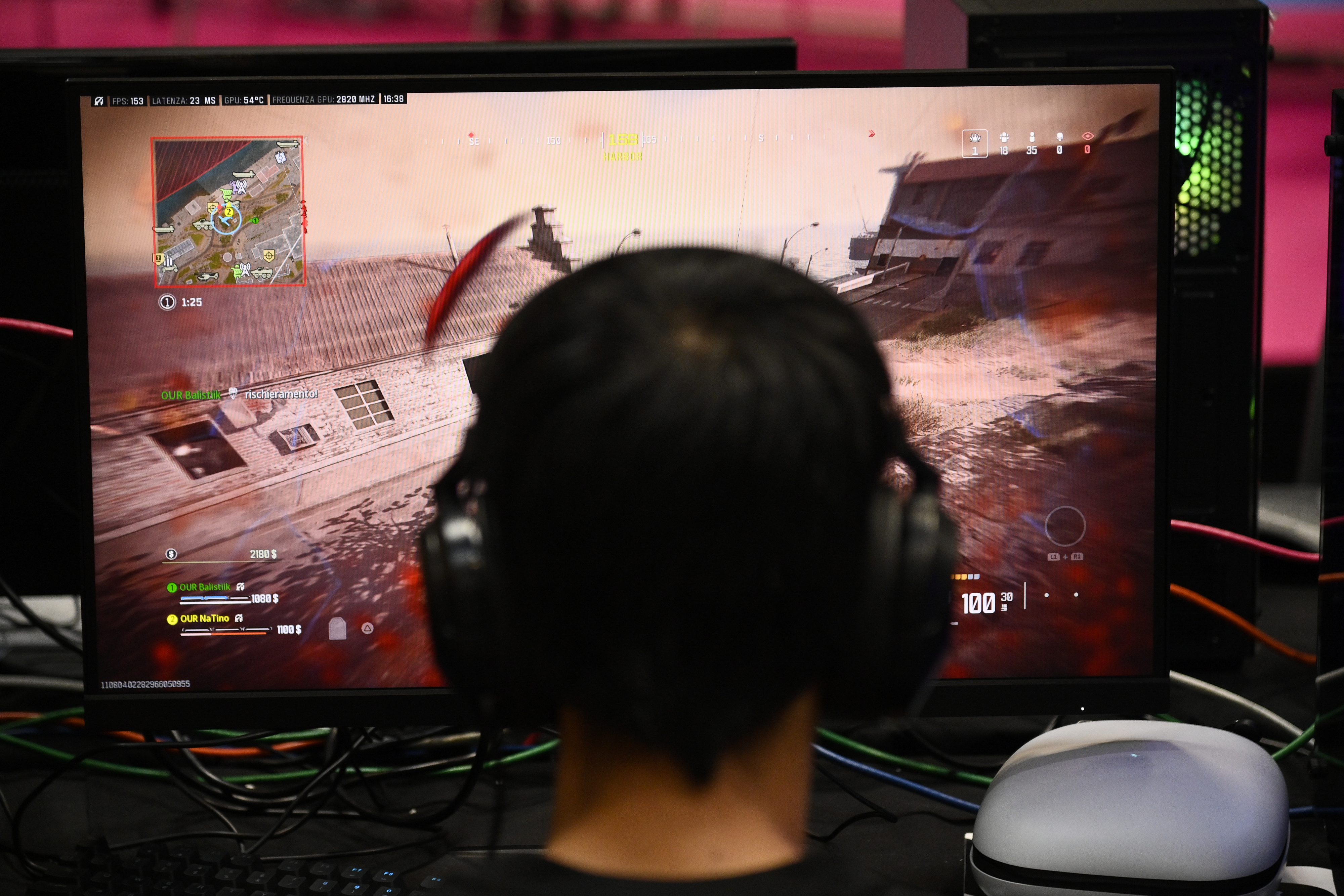 Person with headphones playing a first-person shooter video game on a large monitor at a gaming event. The game shows an action scene and map inset