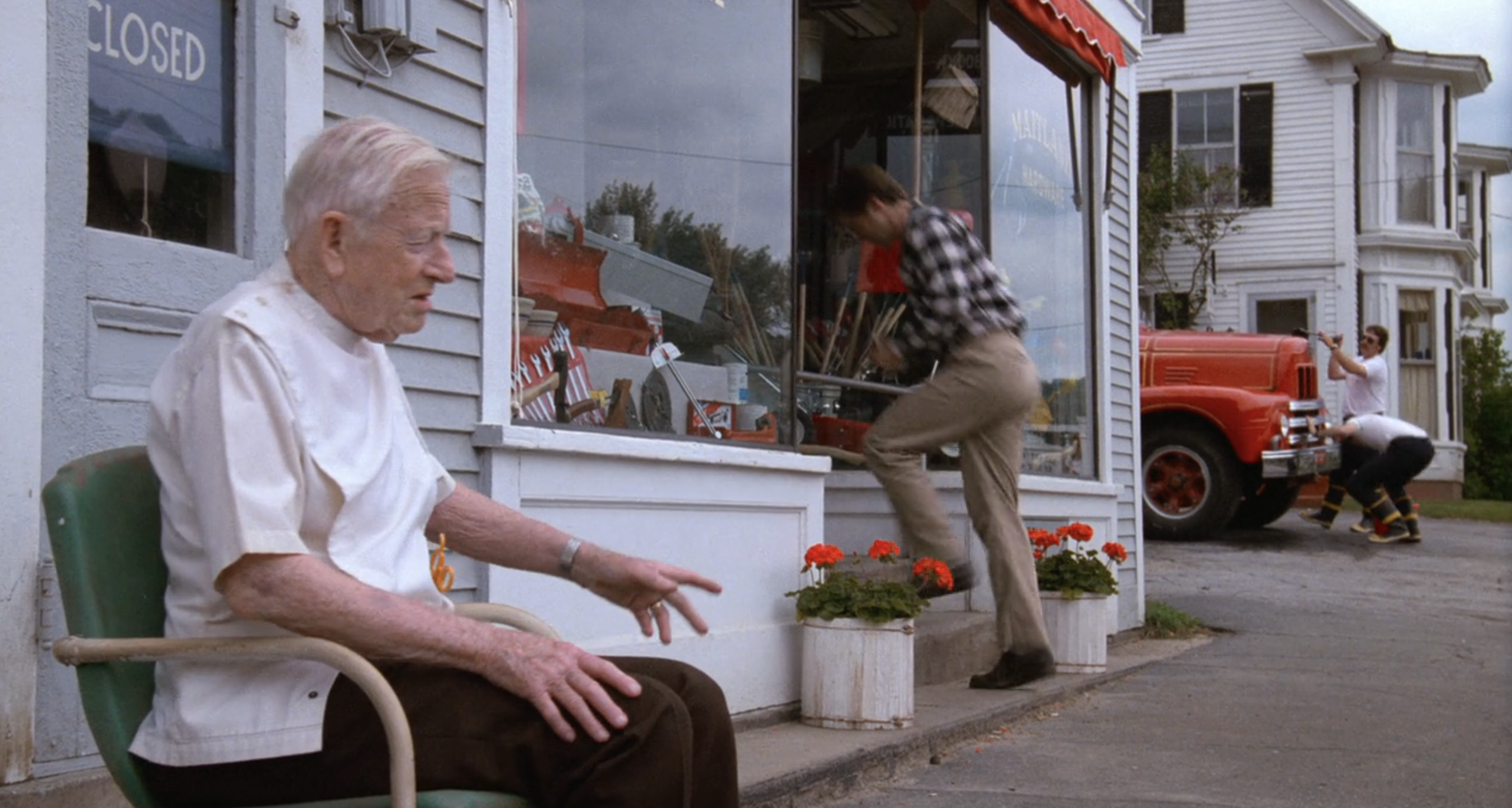 An elderly person sits outside a store while two firefighters push a fire truck uphill in "Beetlejuice"