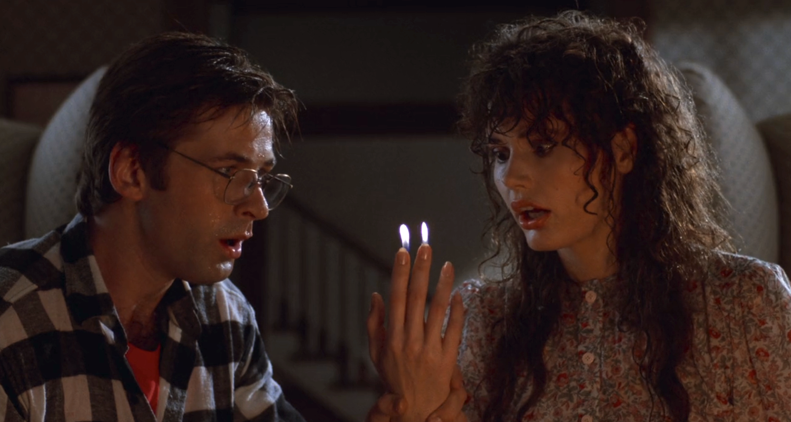 Alec Baldwin with glasses, wearing a plaid shirt, and Geena Davis with curly hair, in a floral blouse, stare amazed at her glowing fingers