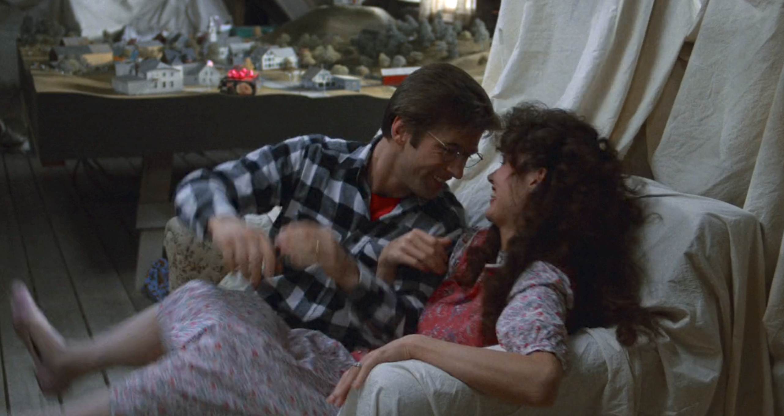 Alec Baldwin and Geena Davis, one in plaid and glasses and one in floral-patterned attire, are joyfully playing or interacting while seated in a room that contains a model village