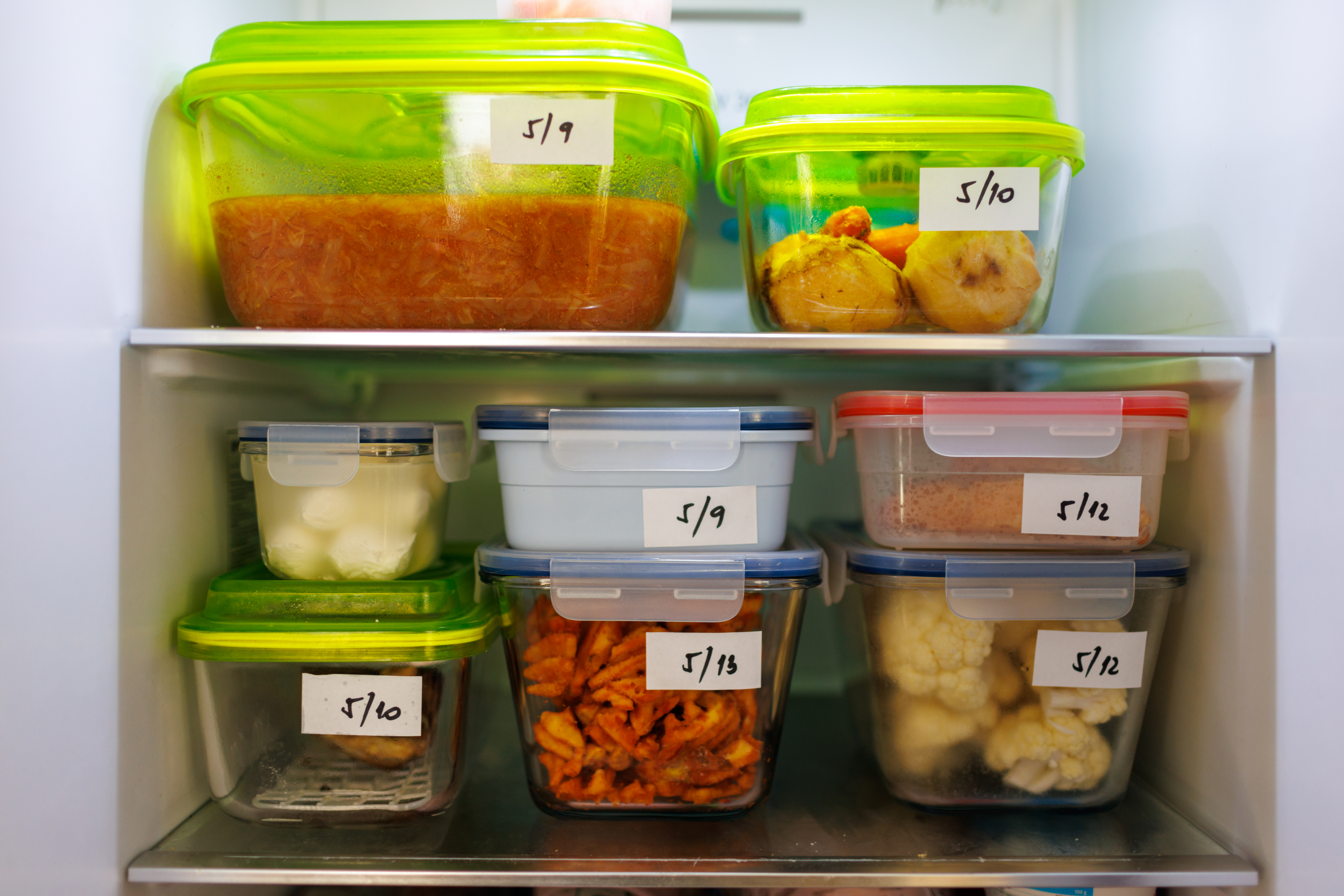Prepared meals and ingredients in labeled containers arranged neatly in a refrigerator. Labels show various dates ranging from 5/9 to 5/15