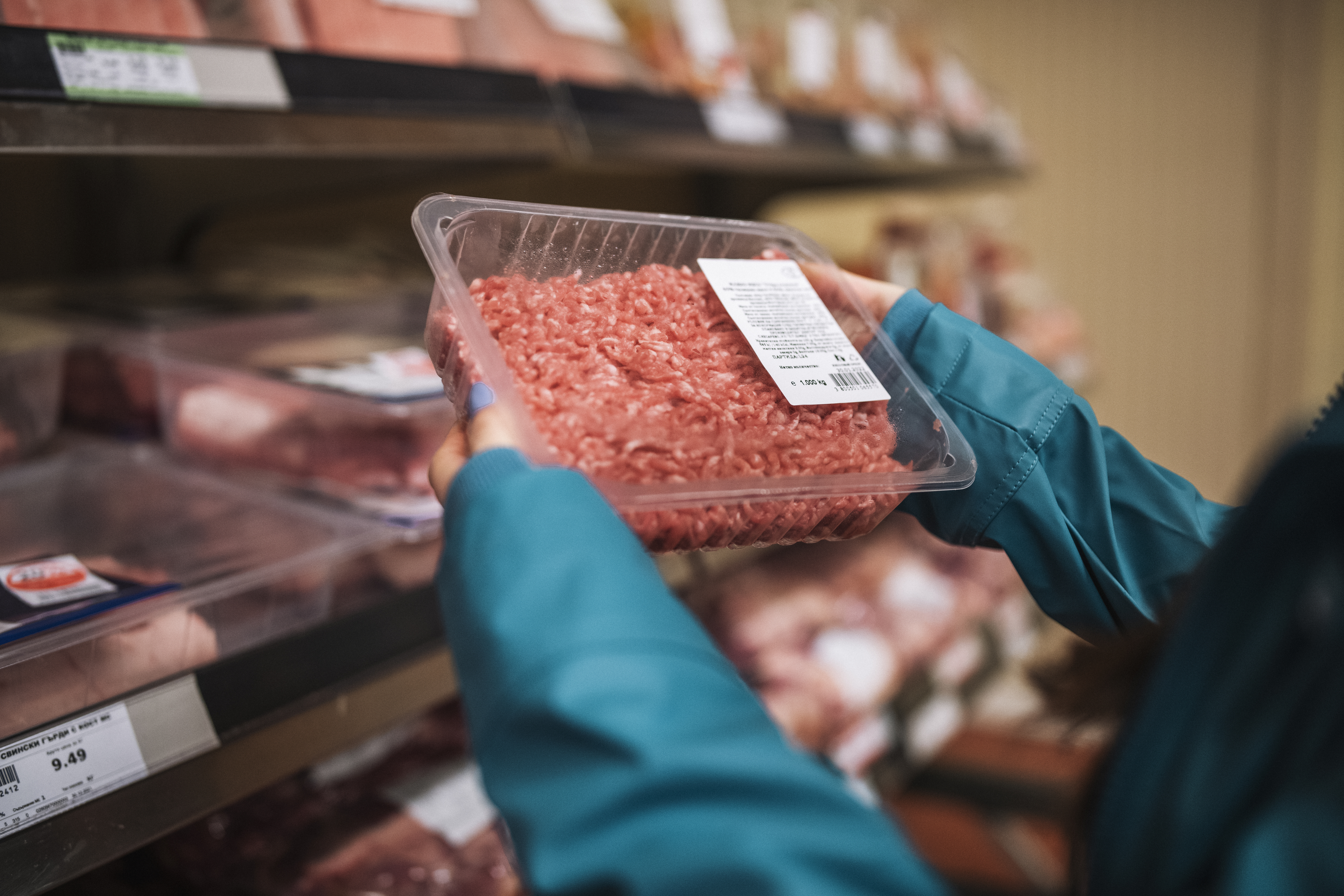 A person holding a packaged tray of ground meat in a supermarket aisle, examining it closely. Shelves with various meat products are in the background