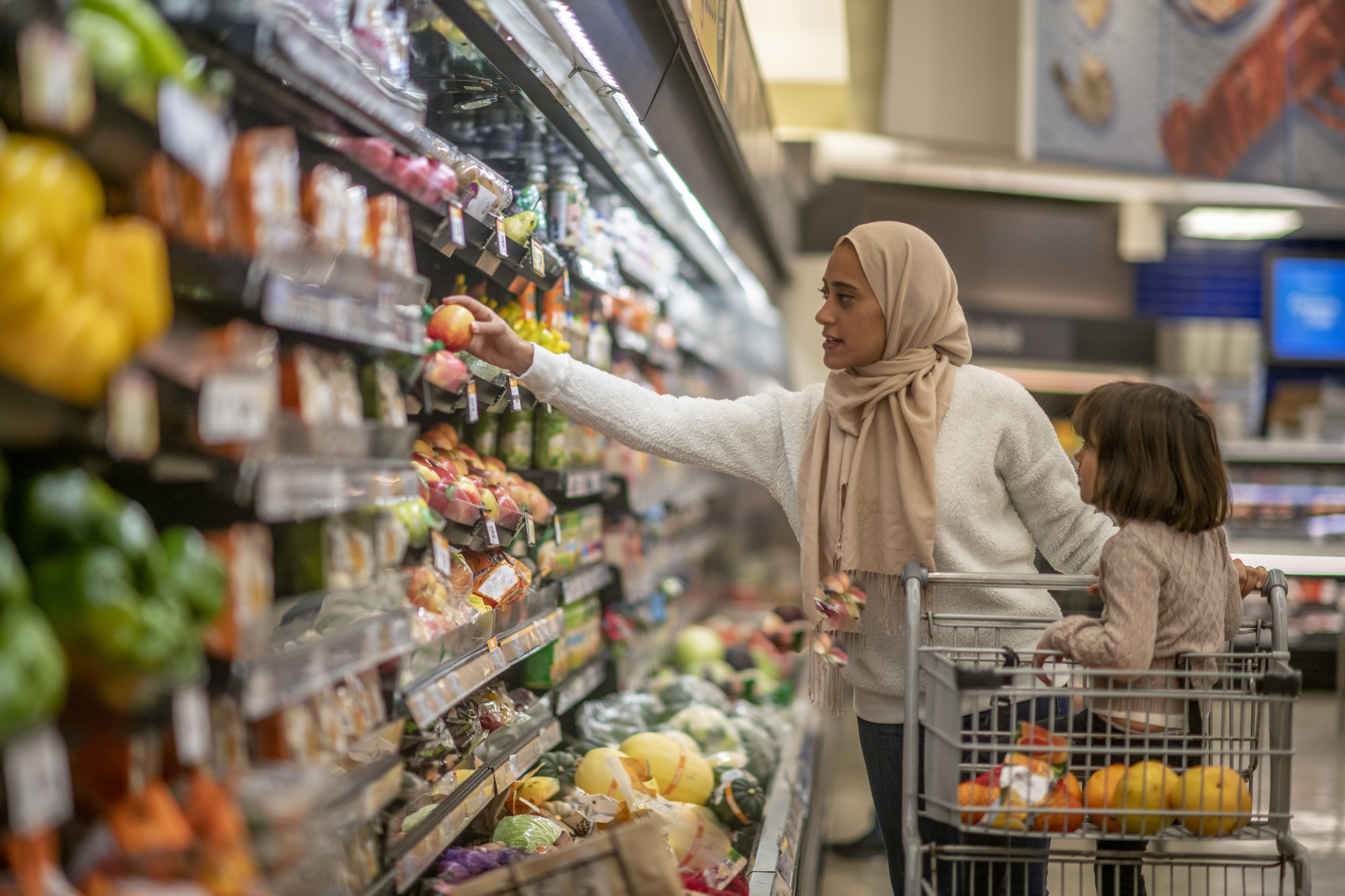 A woman wearing a hijab shops for produce with a young child sitting in a shopping cart in a grocery store
