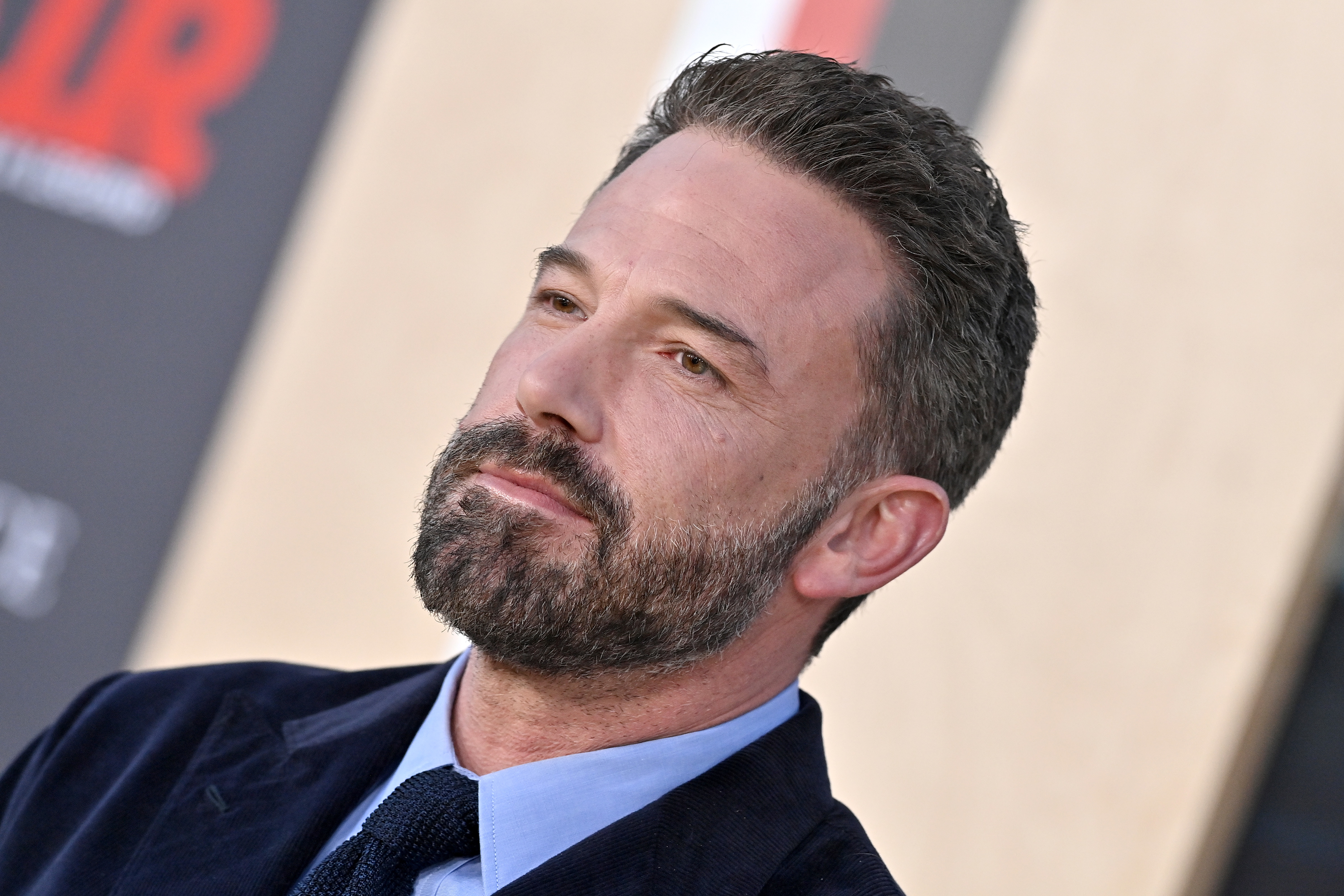 Ben Affleck on a red carpet, wearing a dark suit jacket, blue shirt, and dark tie, looking to the side