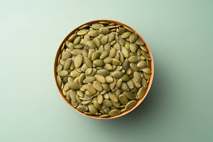 A wooden bowl filled with pumpkin seeds is set against a plain background