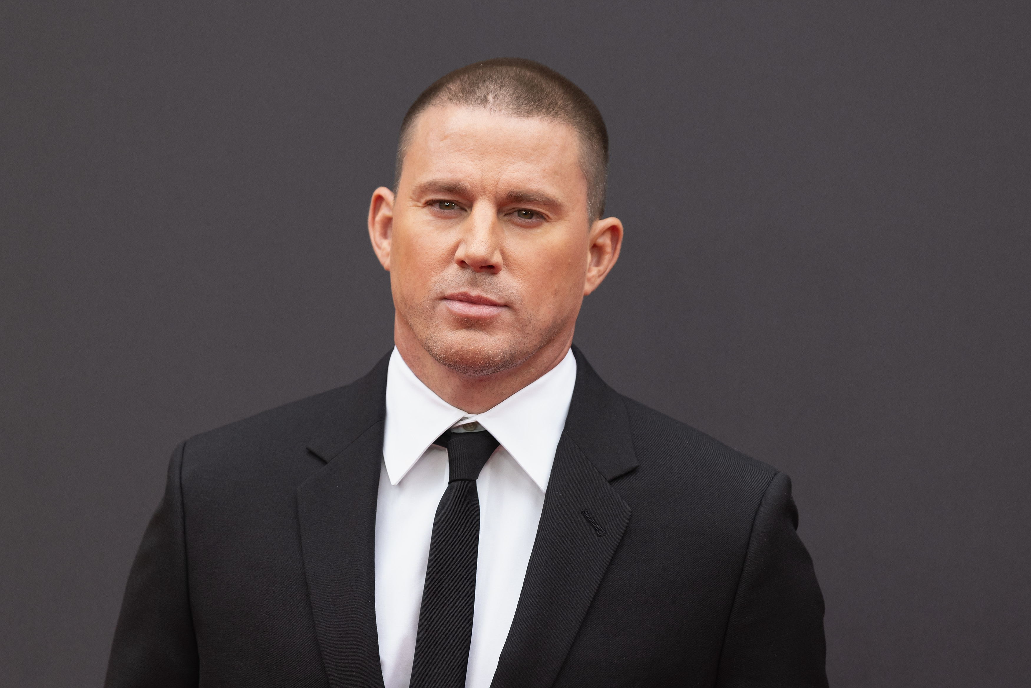 Channing Tatum wearing a black suit with a white shirt and a black tie, posing against a plain background