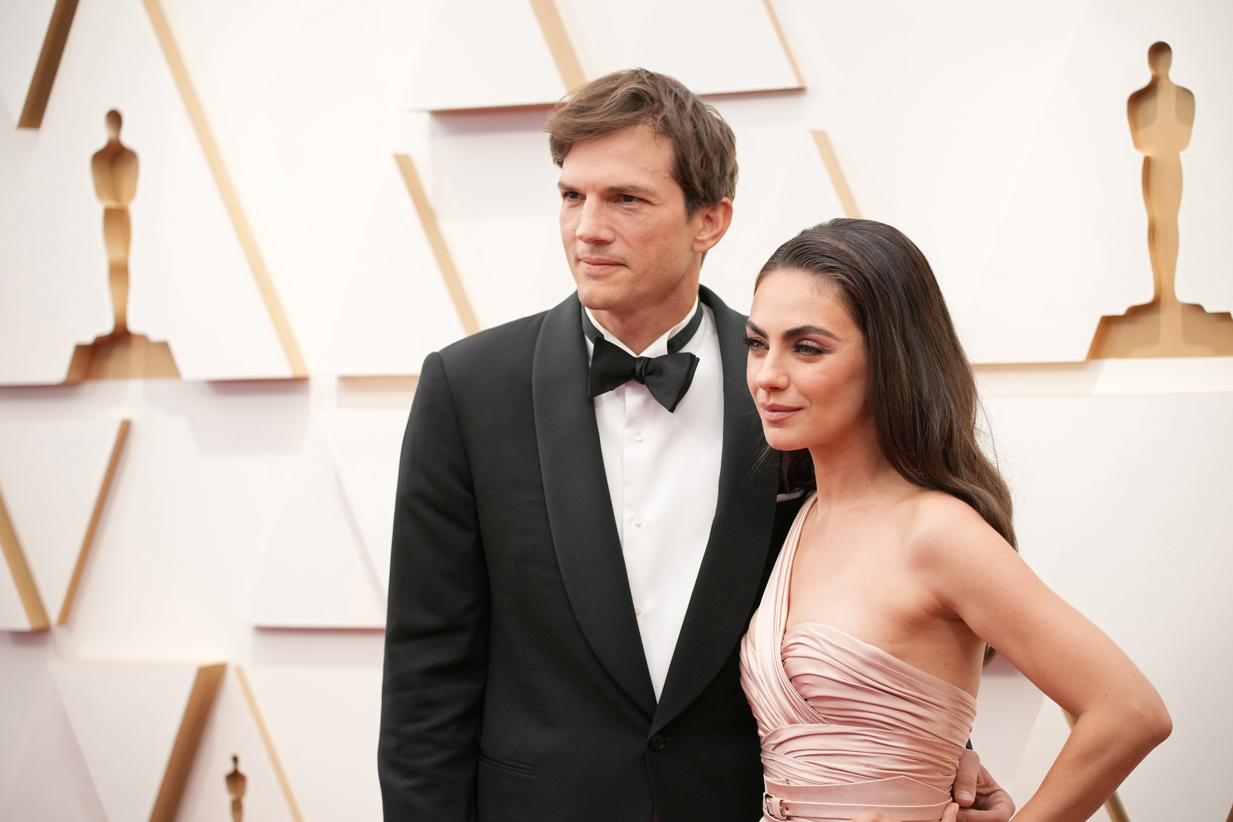 Ashton Kutcher and Mila Kunis on the red carpet. Ashton is wearing a black tuxedo, and Mila is in a stylish off-shoulder gown