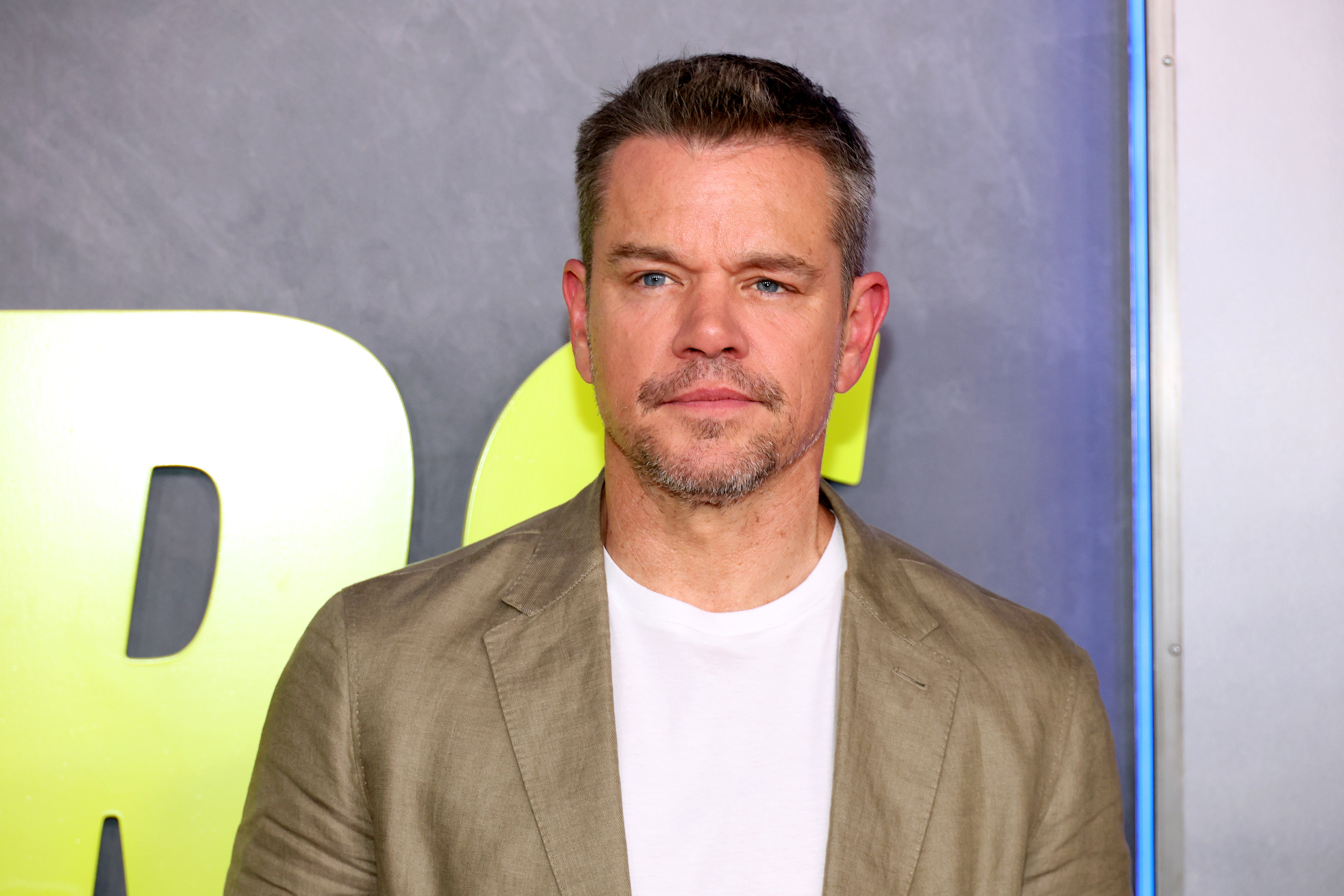 Matt Damon in a casual outfit with a beige blazer over a white t-shirt, posing in front of a backdrop at a media event