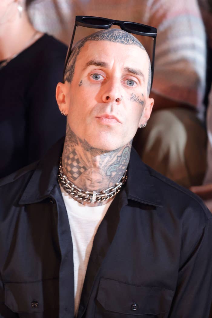 Travis Barker wears a button-up shirt, undershirt, heavy chain necklaces, and sunglasses on his head, showcasing his extensive tattoos