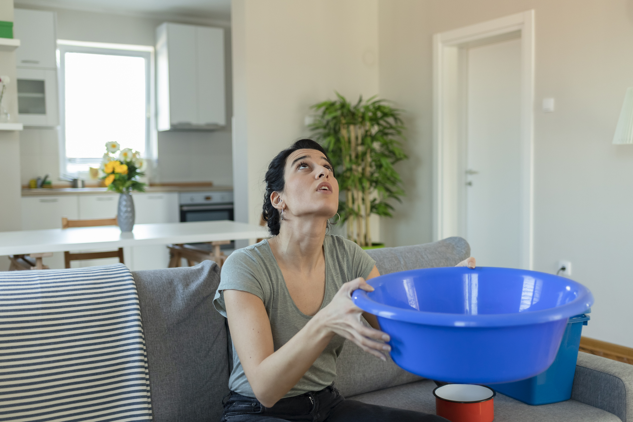 A woman sits on a couch in a living room holding a blue plastic basin, looking up, seemingly concerned about something above her