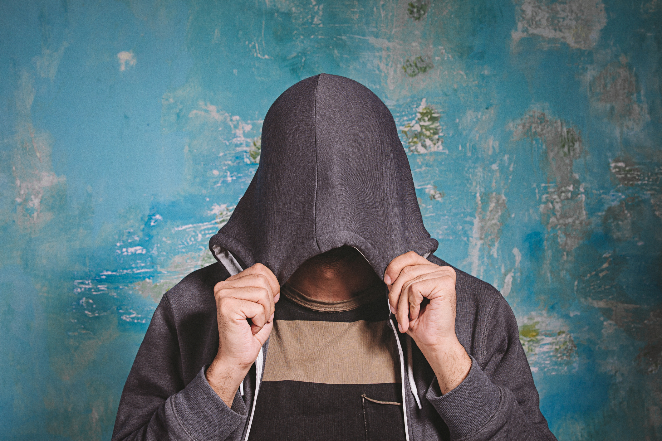 Person wearing a hoodie with the hood covering their face, standing in front of a textured blue background. Face is not visible