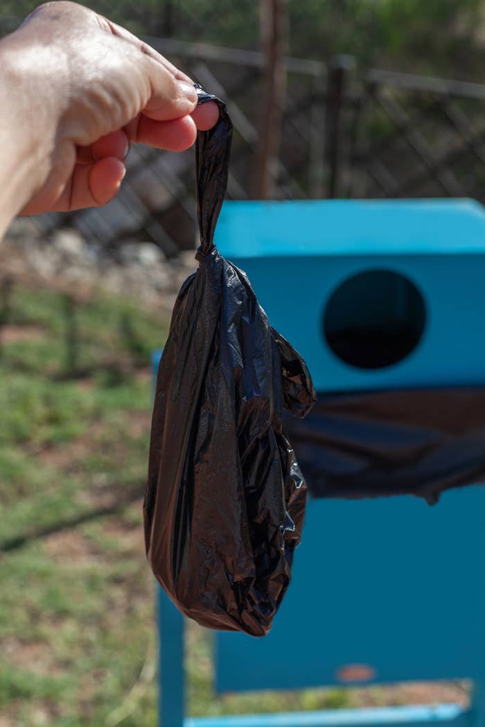 A hand holding a tied black plastic bag near a blue trash bin in an outdoor setting