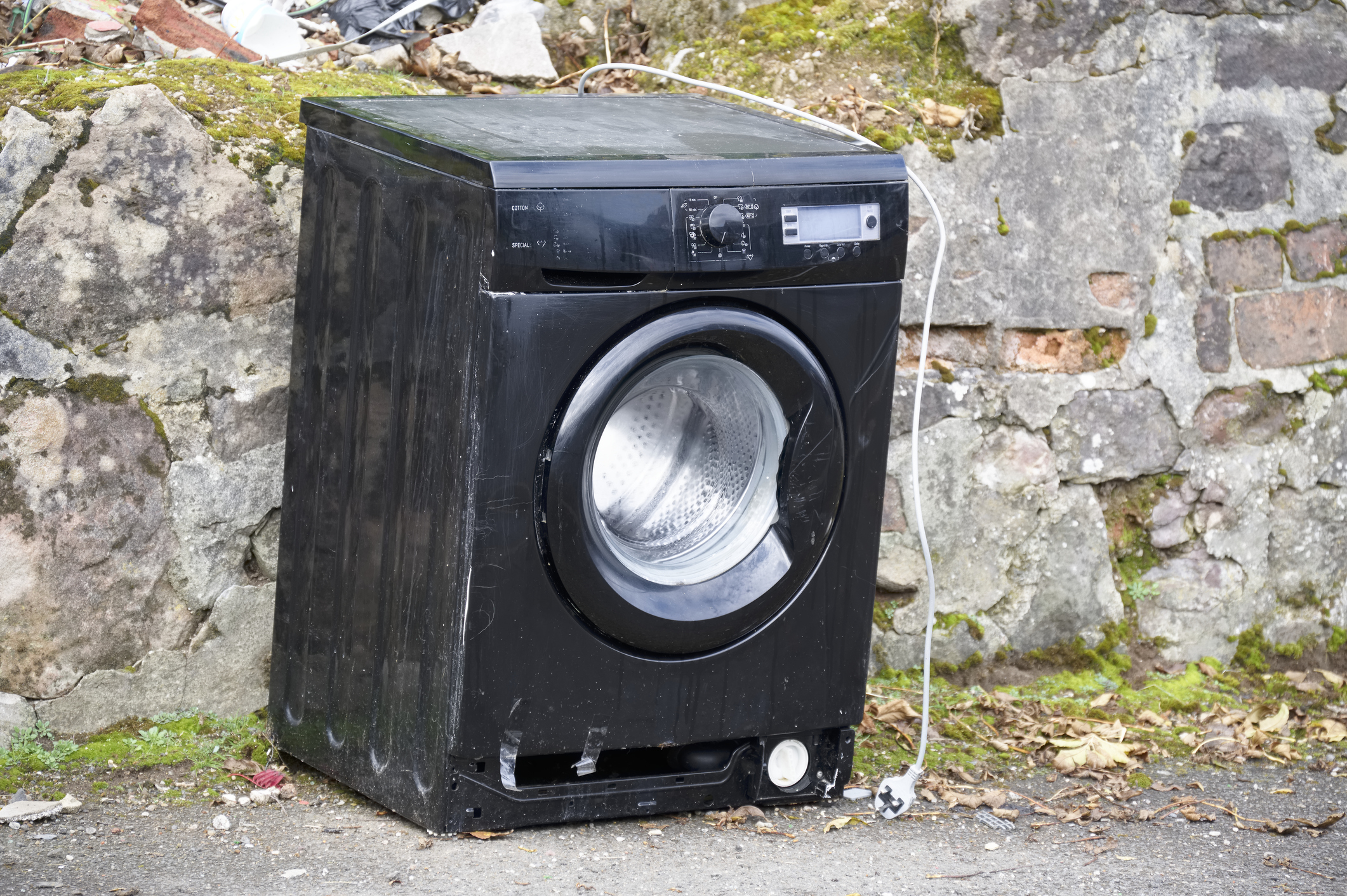 An old, black washing machine is placed outside near a stone wall, with visible signs of wear and tear