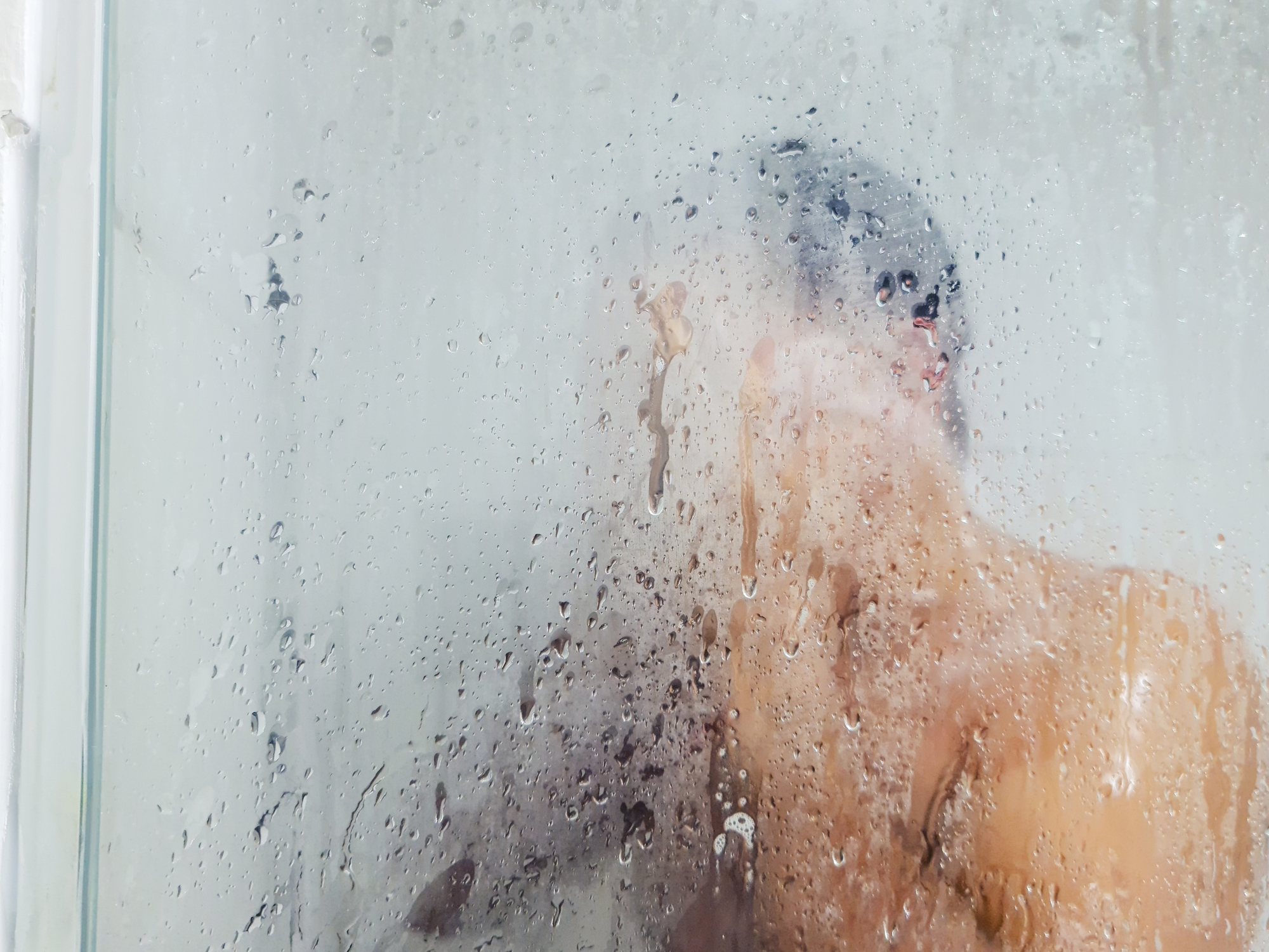 A person is taking a shower behind a foggy glass shower door, with only their upper body partially visible. The image obscures specific details
