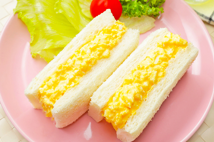 ピンクの皿にのった卵サンドイッチと、レタス、プチトマト、パセリの付け合わせ。