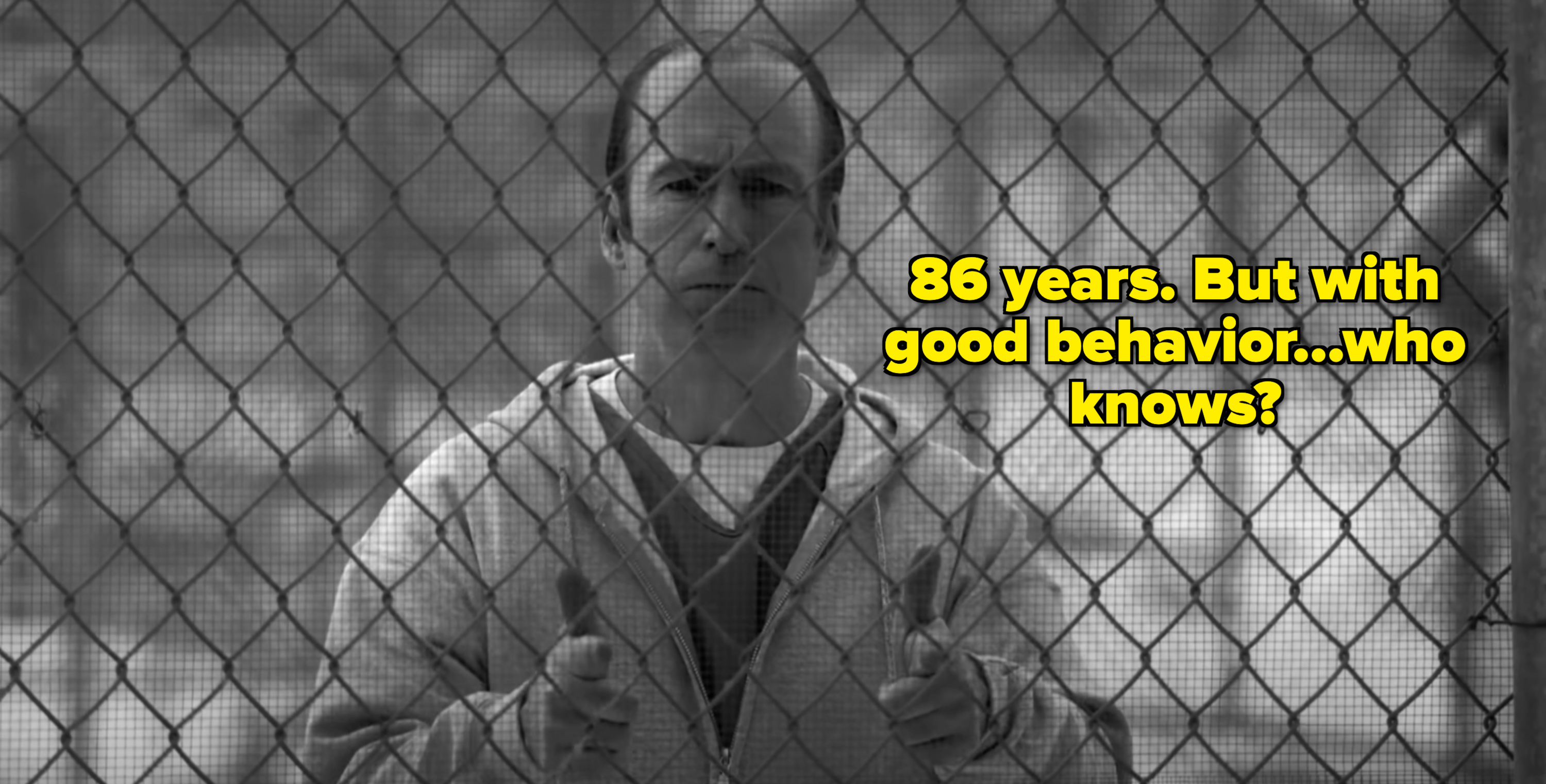 Bob Odenkirk, in character, stands gripping a chain-link fence while wearing a hoodie and looking determined