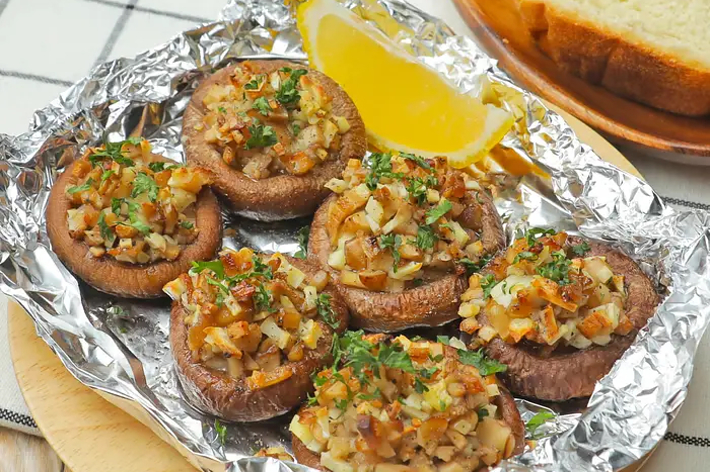 Stuffed mushrooms with a chopped garnish, served on foil with a lemon wedge, and a side of bread on a checkered cloth