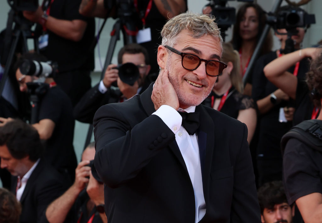 Joaquin Phoenix poses on the red carpet, wearing a tuxedo and bowtie, touching the back of his neck with one hand, with photographers in the background