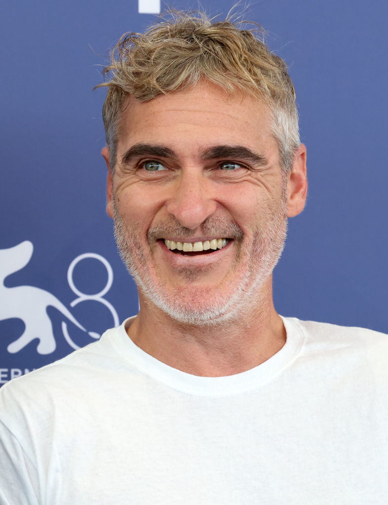 Joaquin Phoenix at a media event, smiling, wearing a casual white t-shirt, with a blue background behind him
