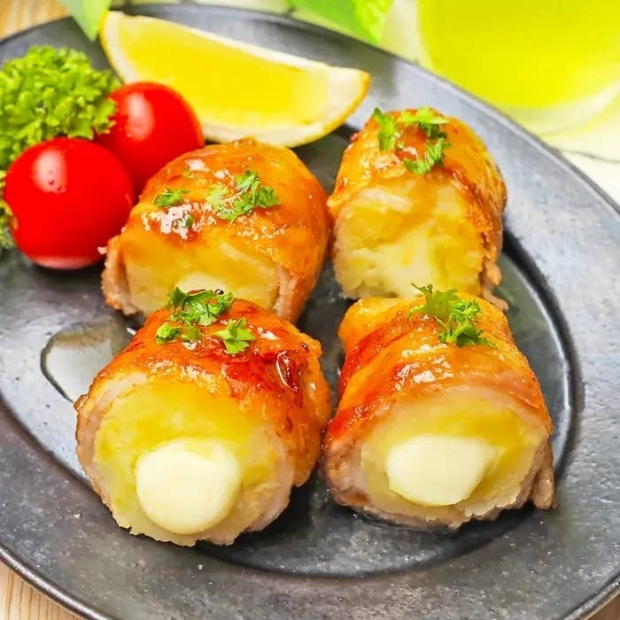 黒いプレートに盛られた豚バラ肉で巻かれたチーズ入りジャガイモの料理。トマトとパセリ、レモンも添えられている。