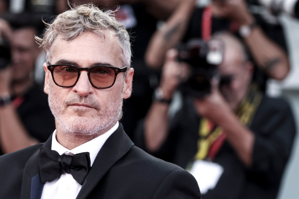 Joaquin Phoenix on a red carpet wearing a black suit with a bow tie. Photographers with cameras are visible in the background