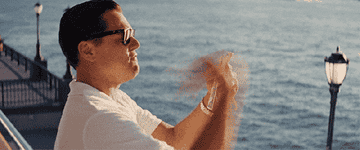 Leonardo DiCaprio clapping enthusiastically while wearing sunglasses and a white shirt on a pier with a body of water in the background