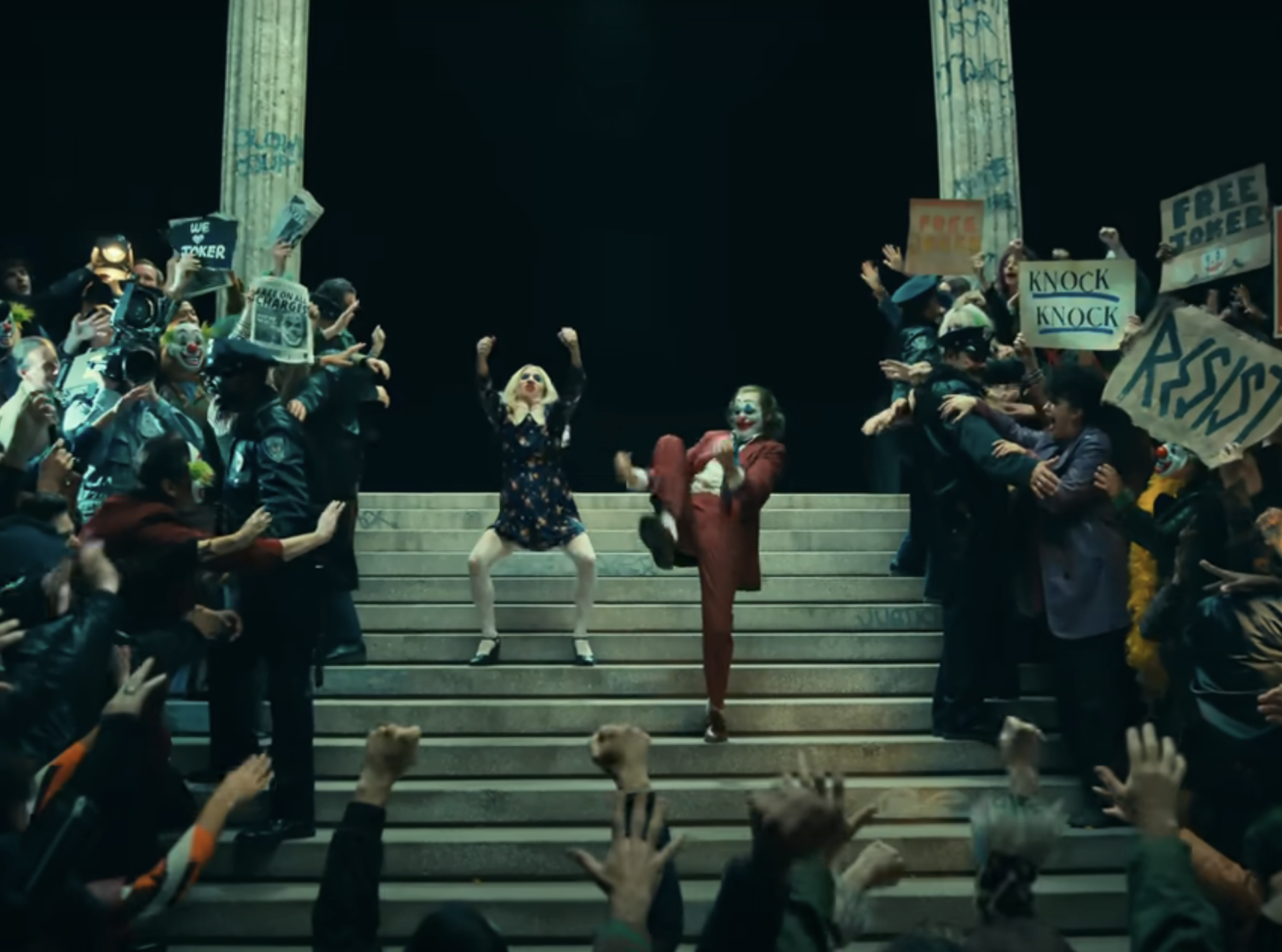 Joaquin Phoenix, in a red suit, and Lady Gaga, in a dress, dance on stairs surrounded by cheering and sign-holding crowd in a protest scene