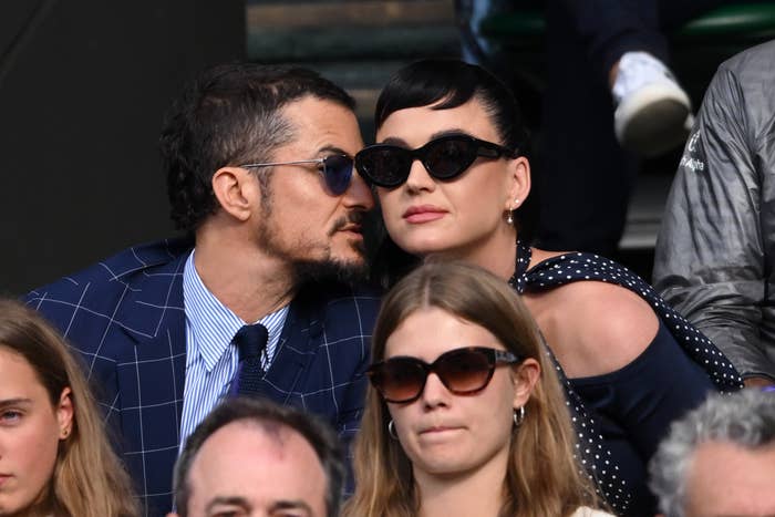 Orlando Bloom and Katy Perry sit closely together at an outdoor event. Bloom wears a checkered suit, and Perry dons cat-eye sunglasses and a polka dot dress