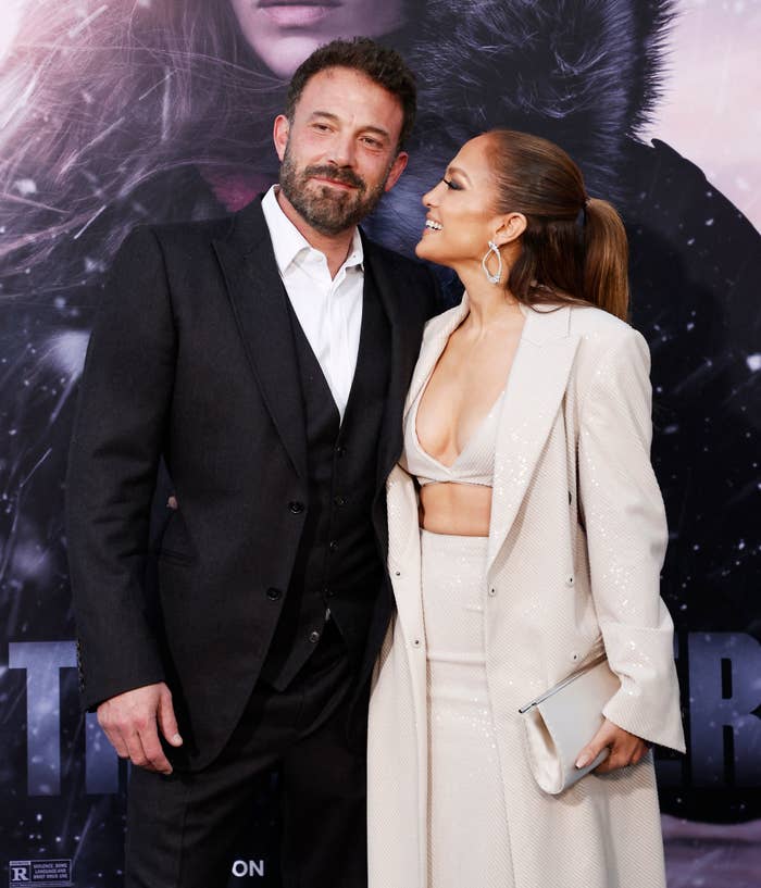 Ben Affleck and Jennifer Lopez posing together at an event. Ben is wearing a suit, and Jennifer is wearing a stylish two-piece outfit with a long coat