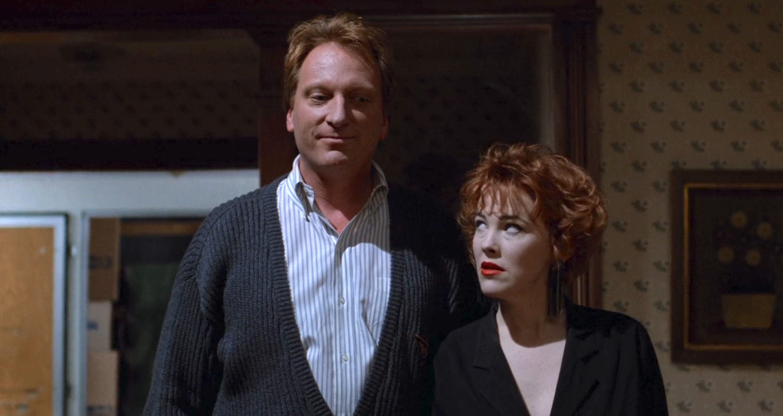 Jeffery Jones wearing a cardigan and Catherin O'Hara with red hair, makeup, and a dark outfit are standing together