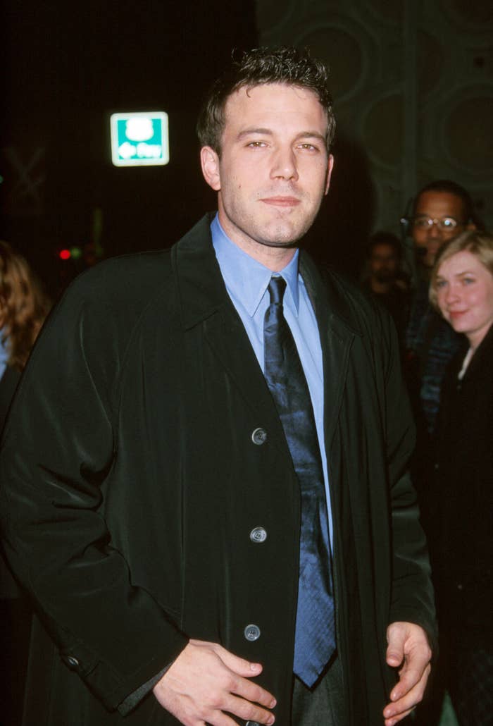 Ben Affleck poses in a dark overcoat and blue tie at an event. The background features other attendees and a green exit sign