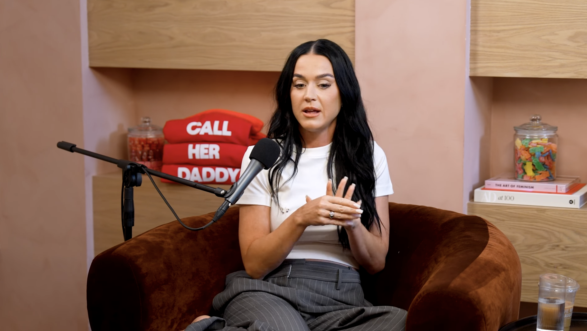 Katy Perry speaks into a microphone during an appearance on the "Call Her Daddy" podcast, sitting in a brown chair with books and jars in the background