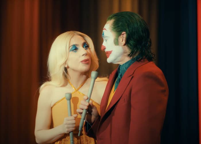 Lady Gaga and Joaquin Phoenix, in character as Harley Quinn and Joker, holding old-fashioned microphones while standing closely together, looking at each other