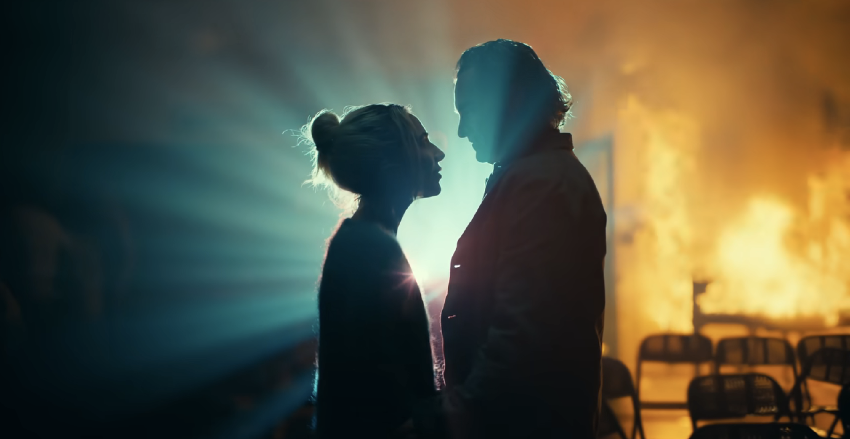 Lady Gaga and Joaquin Phoenix in silhouette, standing close together, against a backdrop of bright lights and fire