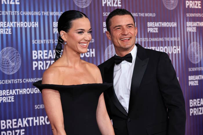 Katy Perry, in an off-the-shoulder dress, and Orlando Bloom, in a tuxedo, smiling on the red carpet at the Breakthrough Prize event