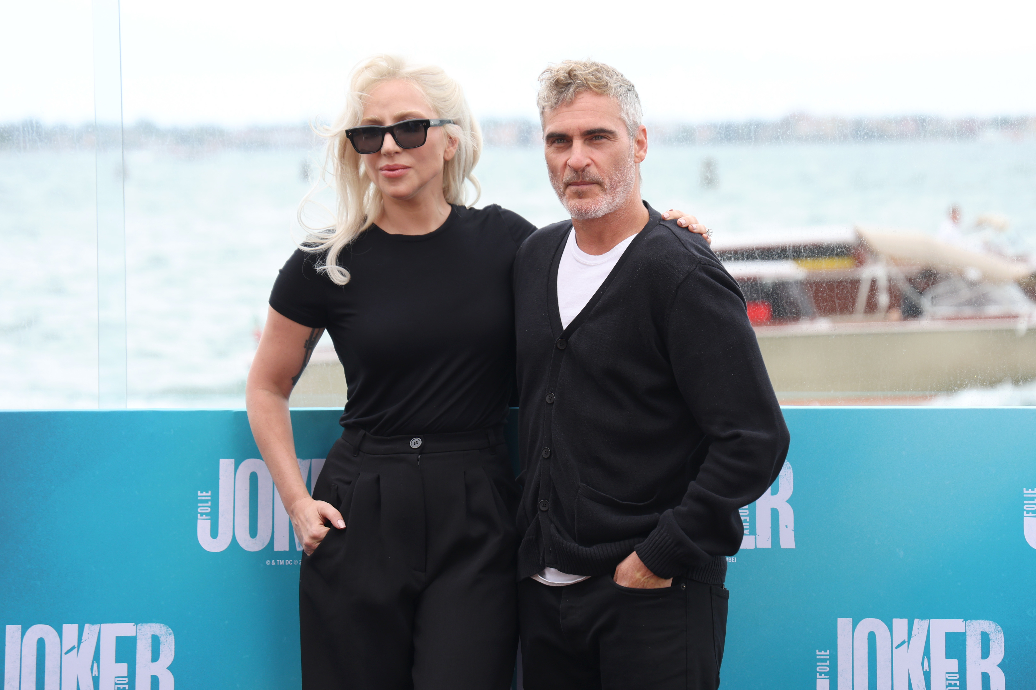 Lady Gaga and Joaquin Phoenix pose together in front of a waterfront backdrop at a promotional event for &quot;Joker.&quot; Gaga wears a black, fitted outfit with sunglasses