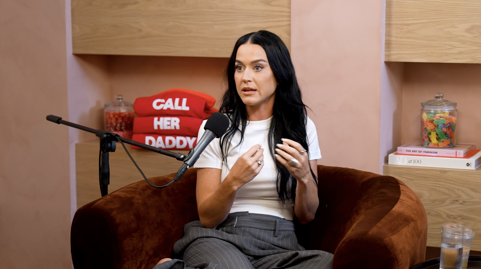 Katy Perry wearing a white top and gray pants, sits on a brown chair during a podcast interview, red "Call Her Daddy" pillows in the background