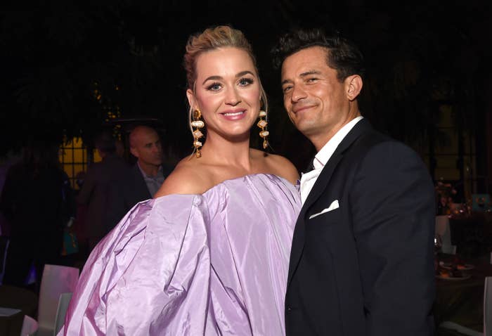 Katy Perry and Orlando Bloom pose together at an event. Katy wears an off-the-shoulder gown and large dangling earrings, and Orlando wears a dark suit with a white shirt