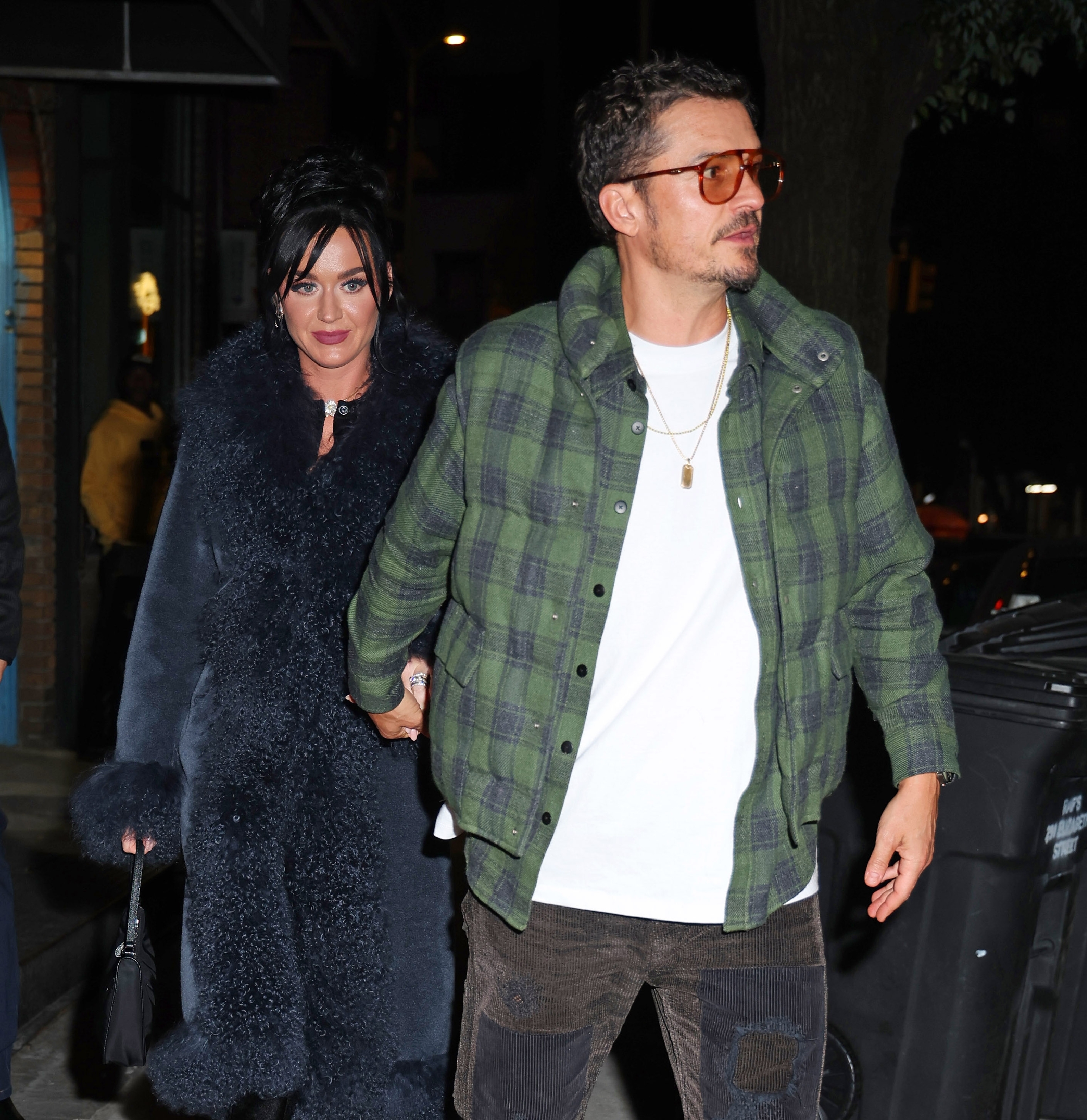 Katy Perry and Orlando Bloom walking together, holding hands. Katy is wearing a long, fur coat, and Orlando is in a plaid shirt with sunglasses