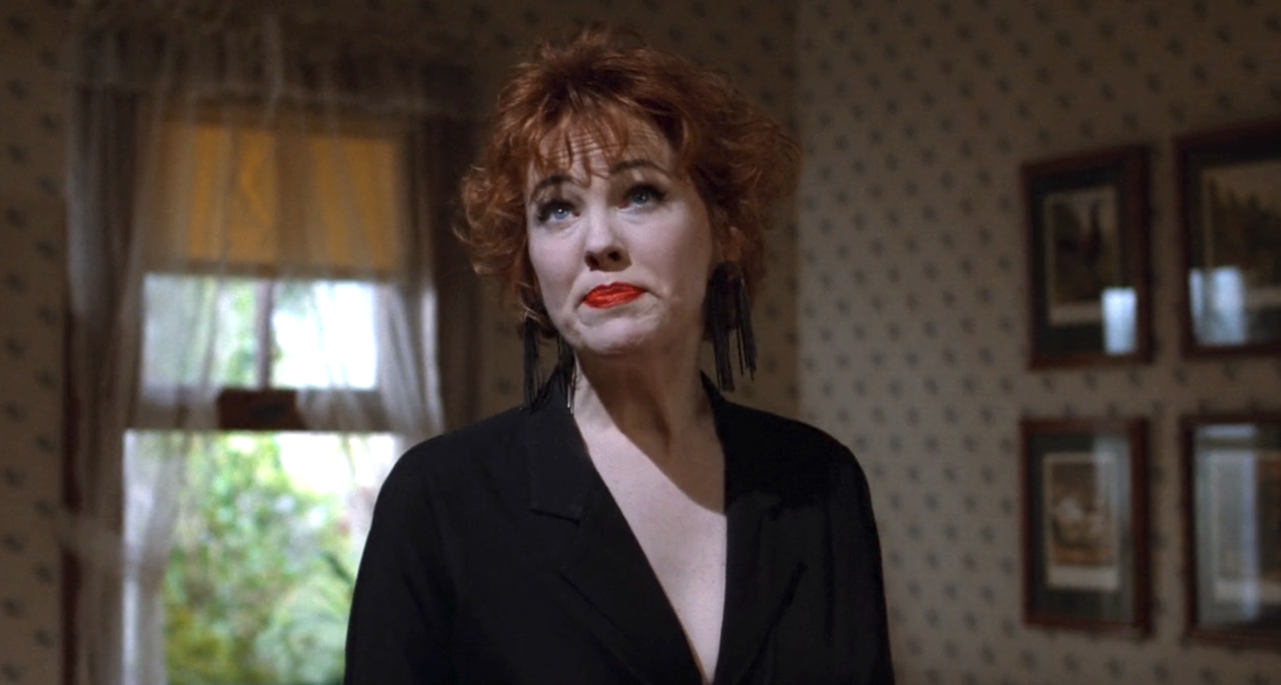 Catherine O'Hara with red hair, earrings, and a dark V-neck top stands indoors, looking upward