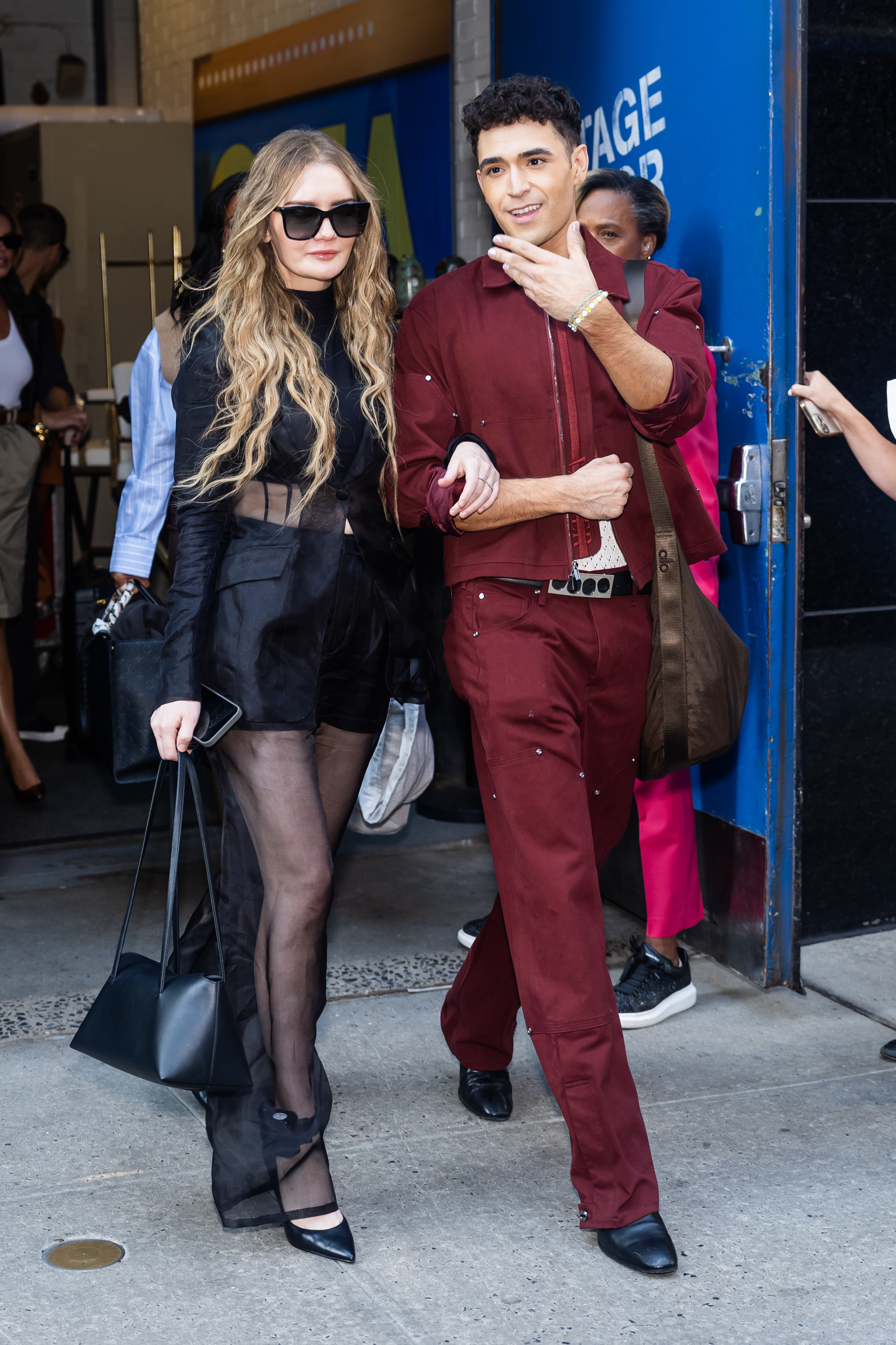 Anna Delvey and Richie Shazam exit a building, with Julia in sheer black outfit and Richie in a red outfit. Julia wears large sunglasses and carries a black bag