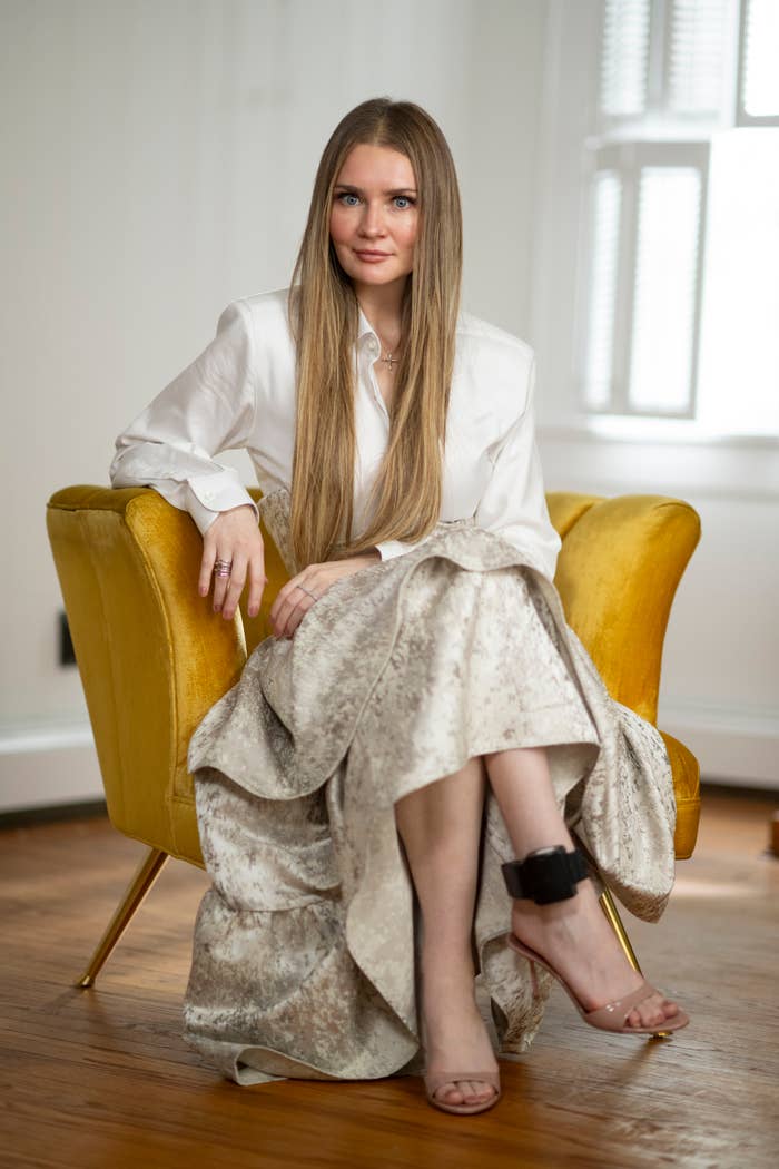 Anna Sorokin sits on a yellow chair, wearing a long blouse and an ankle monitor