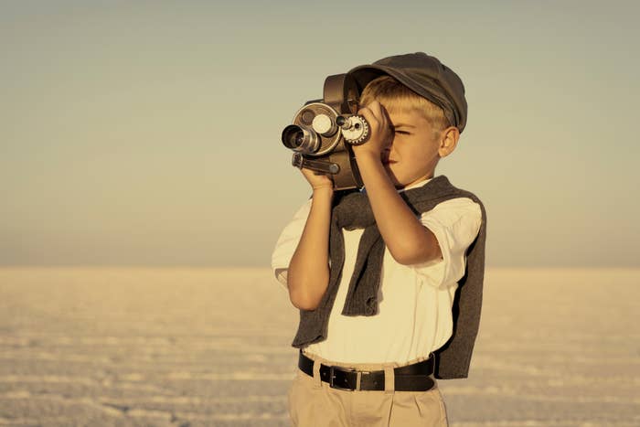 A young boy in vintage attire, wearing a cap and holding an old-fashioned movie camera, looks through the viewfinder while standing in a vast, barren landscape