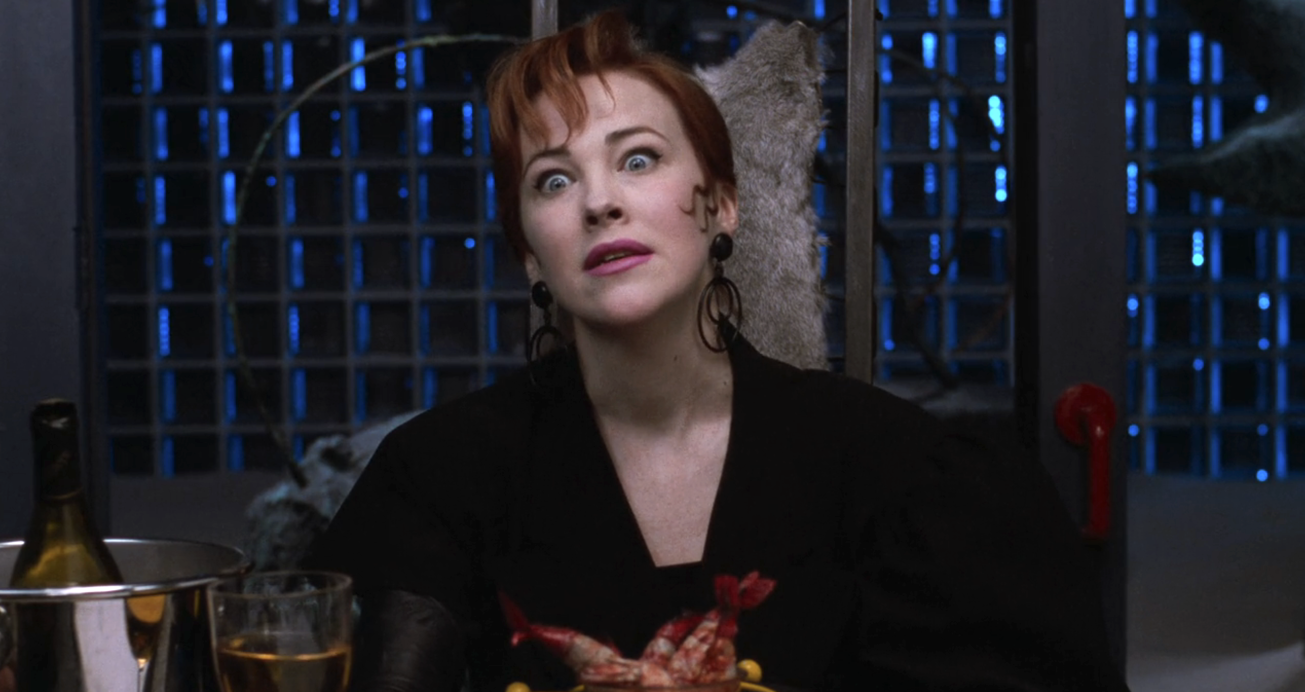 Catherine O'Hara is sitting at a table with a surprised expression, with food and drinks in front of her