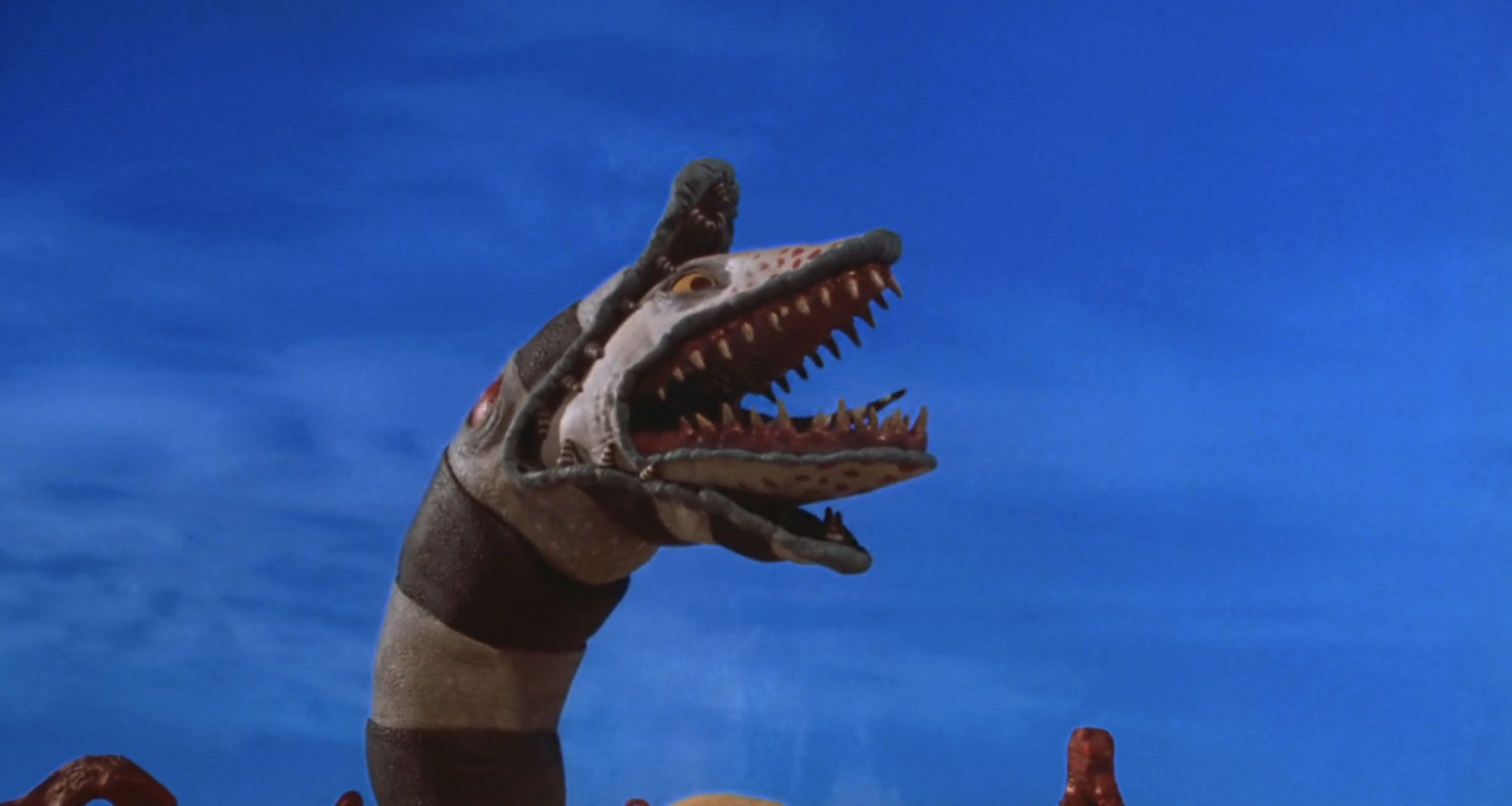 The image shows a large, sandworm-like creature with several mouths open wide against a blue sky background in "Beetlejuice"
