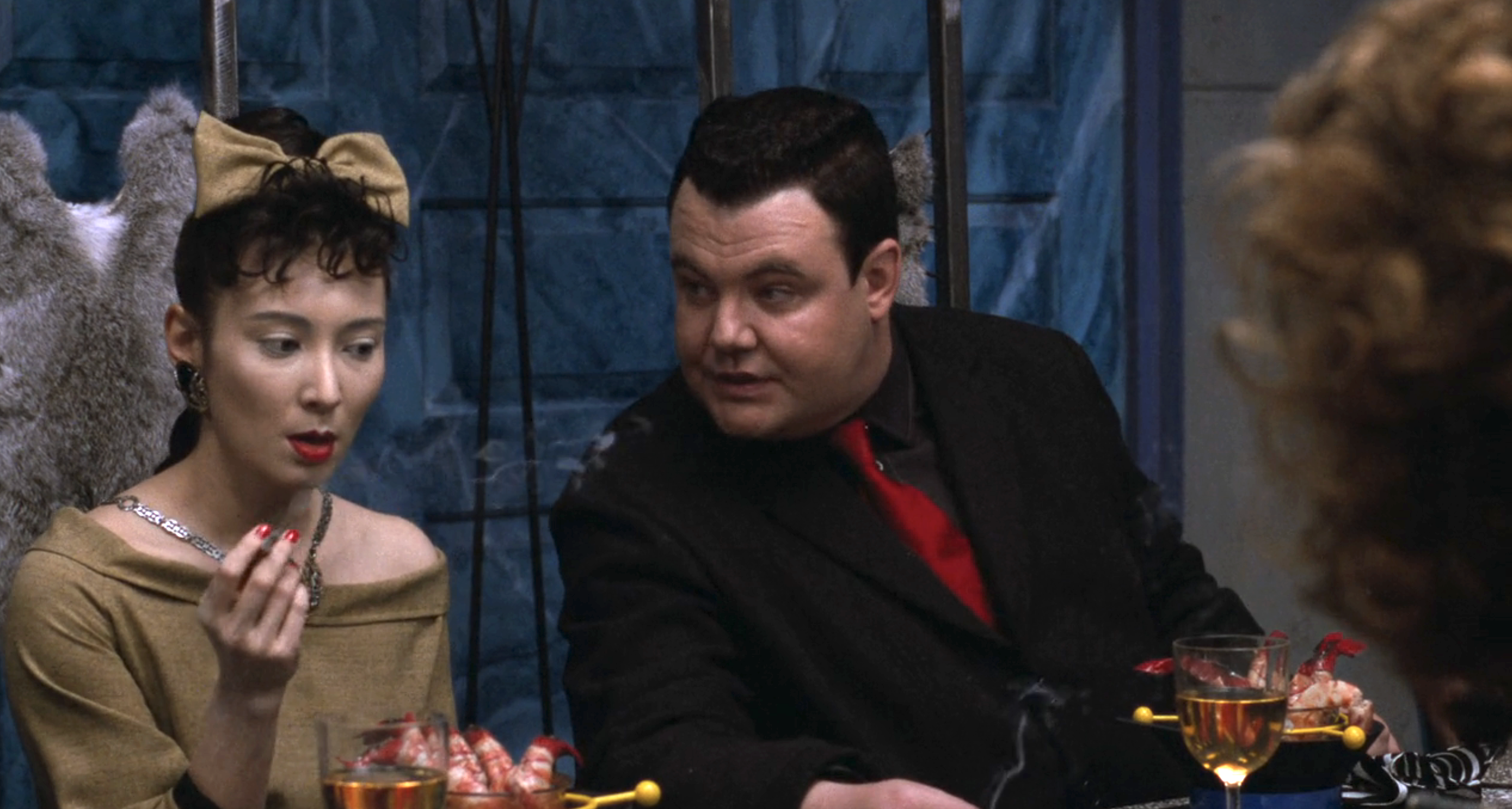 An actress and Glenn Shadix, dressed in formal attire, are seated at a table engaged in conversation, with drinks and shrimp cocktail in front of them