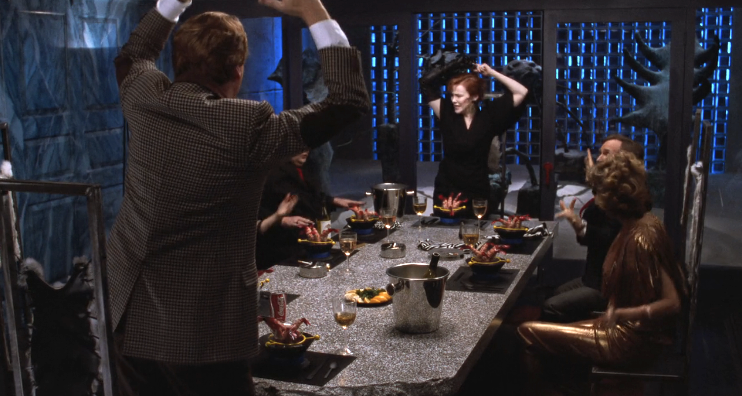 Group of people having dinner at an elegant table. Catherine O'Hara stands with arms raised, while others are seated and smiling. The setting looks theatrical and dramatic