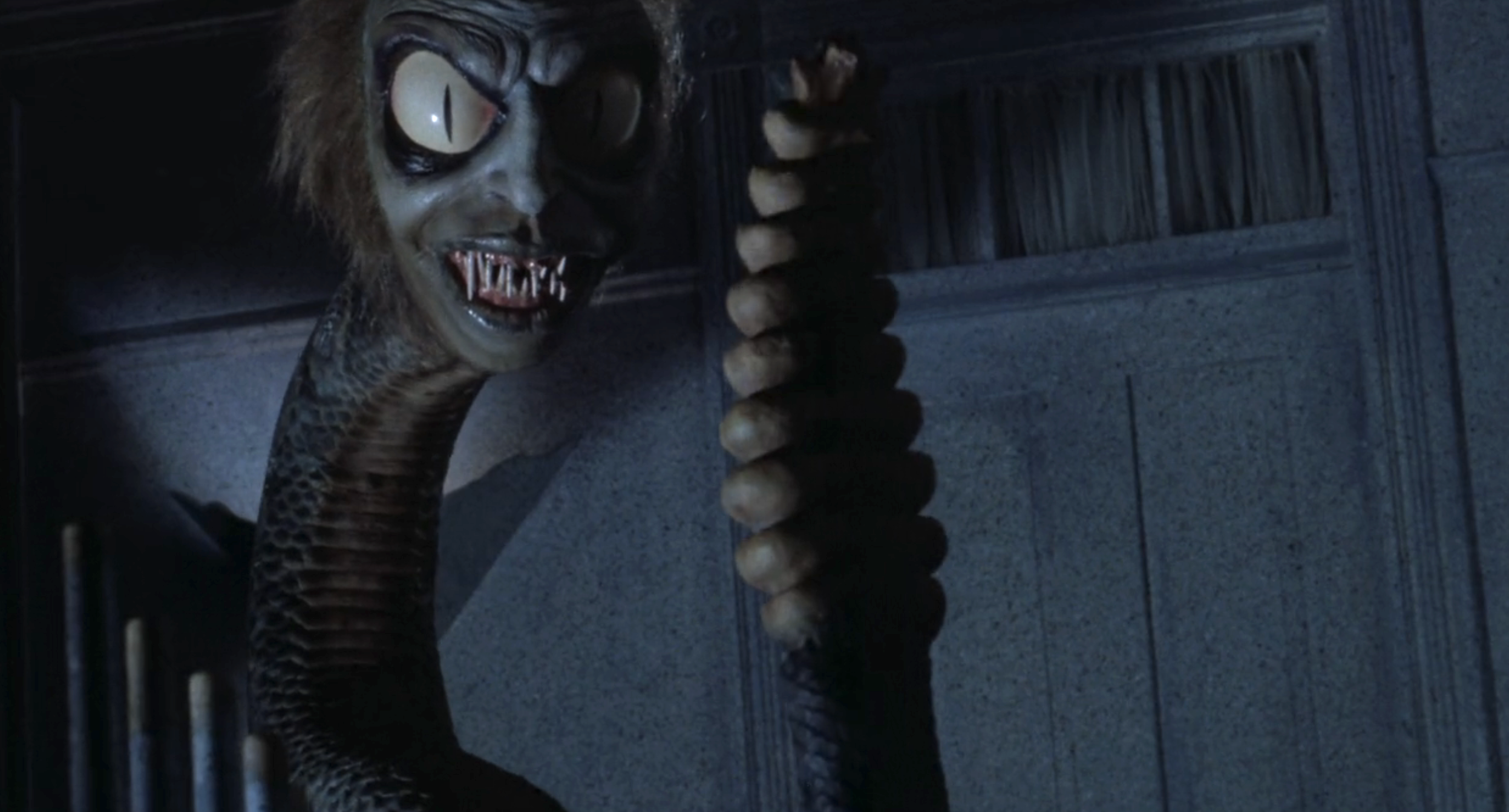 A close-up of a monstrous creature from the movie Beetlejuice, with large eyes, sharp teeth, and a serpent-like body, holding a wooden object